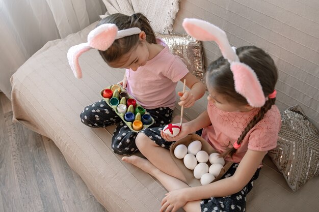 Little sisters in bunny ears paint Easter eggs on the sofa at home