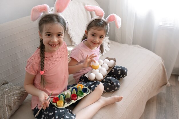 Little sisters in bunny ears paint Easter eggs on the sofa at home