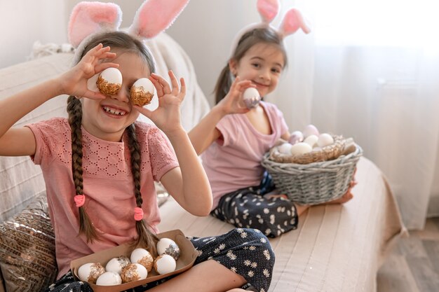 Little sisters with bunny ears, with Easter eggs at home on the couch