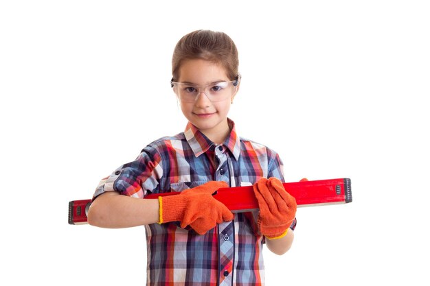 Маленькая улыбающаяся девочка в клетчатой красной рубашке с оранжевыми перчатками и очками, держащая строительный уровень