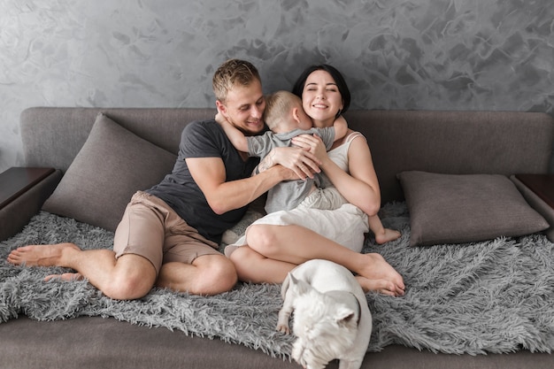 Little son hugging her parents sitting on sofa with white dog