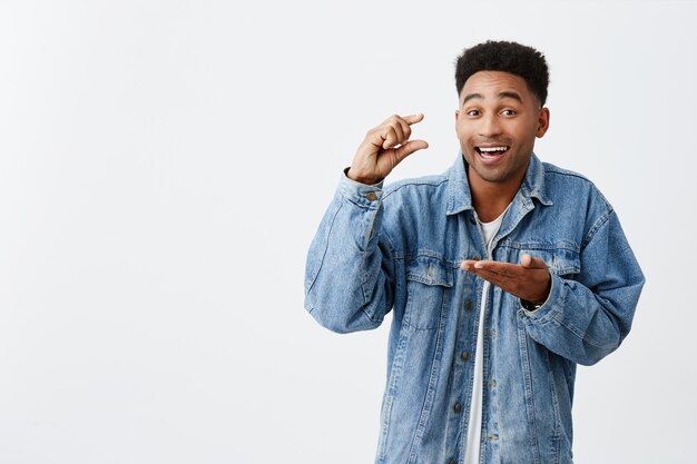 Little. Tiny. Isolated portrait of young good-looking funny happy black-skinned man with afro hairstyle in casual denim jacket gesticulating with hands, looking in camera with joyful expression