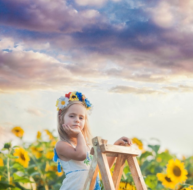 The little ukrainian girl is decorated with a wreath, standing on a wooden ladder among a sunflower field
