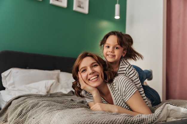 Little woman and her mother are lying on bed, laughing and looking into camera.