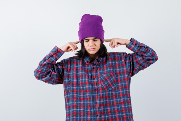 Little woman plugging ears with fingers in checkered shirt and beanie looking annoyed