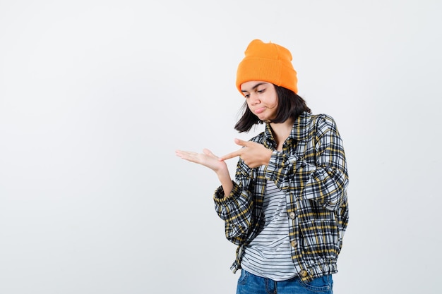 Little woman pointing at palm in t-shirt and jacket and beanie looking hesitant