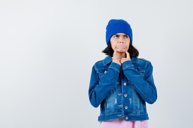 Little woman pulling down cheeks with fingers in t-shirt and denim jacket and beanie