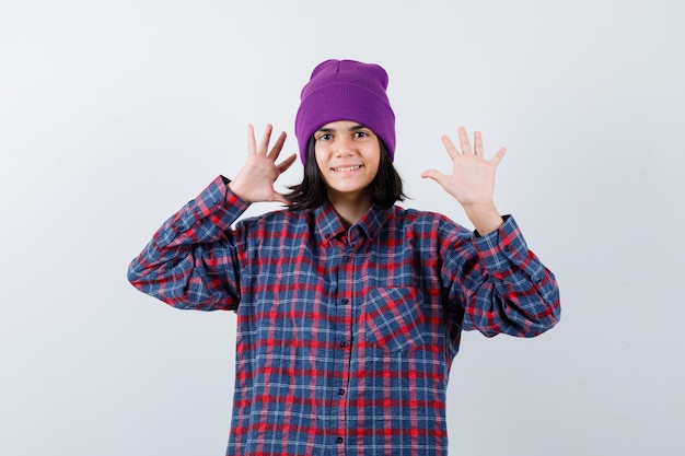 Little woman showing palms in checkered shirt and beanie and looking happy