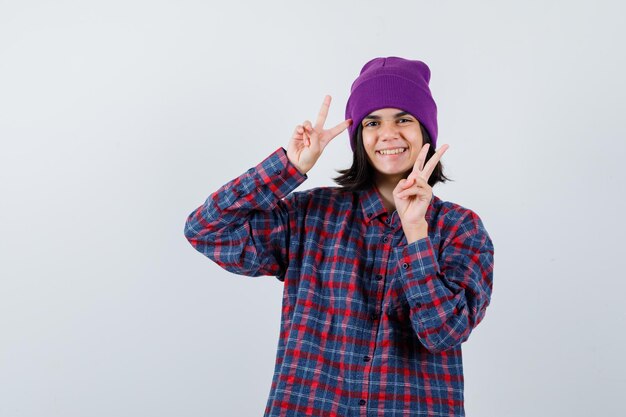 Little woman showing victory sign in checkered shirt and beanie and looking cheery