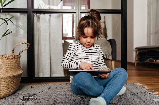 La piccola donna in jeans attillati e maglione a righe disegna in tablet e si siede sul pavimento nel soggiorno.