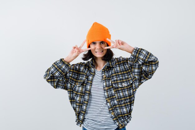 Little woman in t-shirt and jacket and beanie showing victory sign and looking blissful