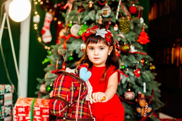 赤いドレスのLittlleの女の子が部屋の緑のクリスマスツリーの前に座っている