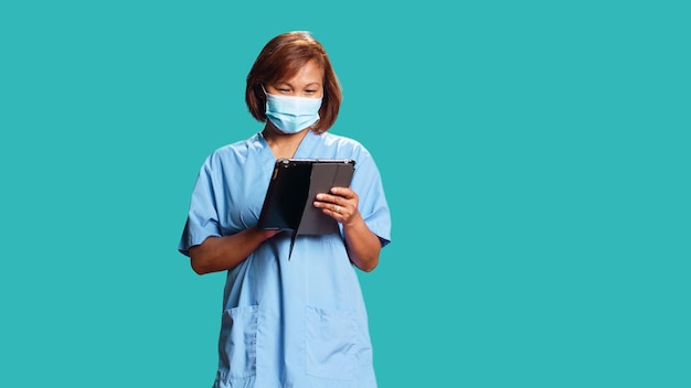 Lively asian nurse looking at patient data on digital tablet, confused with medical checkup results. Professional clinical worker wearing protective face mask, isolated over blue studio background