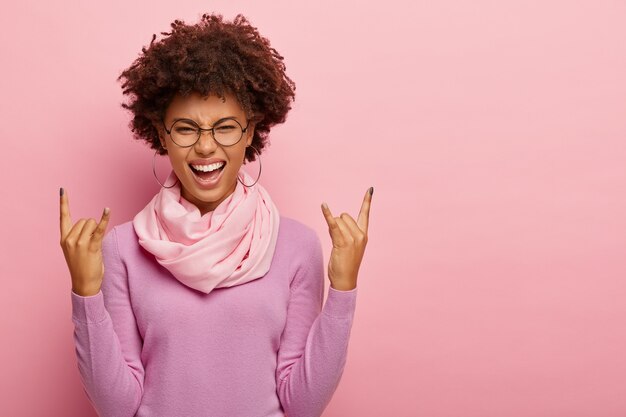 Lively dark skinned young lady makes rock n roll or heavy metal gesture, feels energized, smiles happily, wears glasses and purple poloneck, isolated over pink background