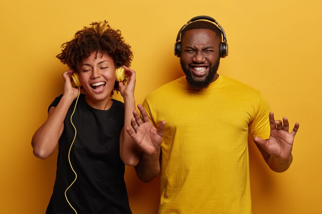 Lively energetic dark skinned couple dance and have fun together, listen different types of music in headphones isolated on bright background.