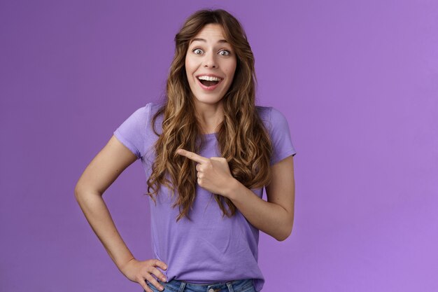 Lively excited enthuasitic good-looking woman curly hairstyle open mouth fascinated look admiration joy share awesome place location pointing left smiling broaldy thrilled purple background