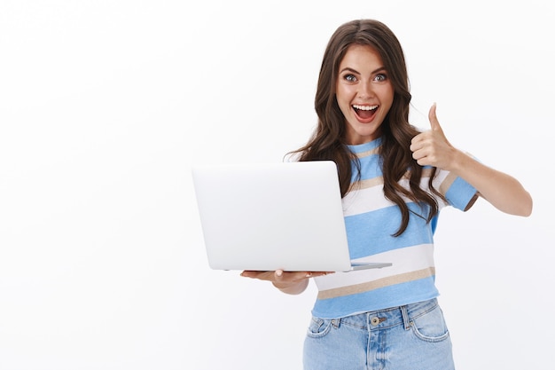 Lively satisfied modern gorgeous woman picked cool computer, smiling broadly hold laptop, show thumb up in approval, accept gesture, like cool friend project music-video, leave positive comment