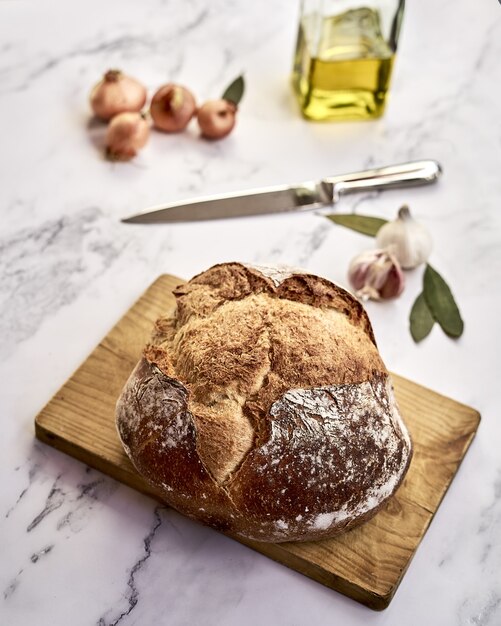 Loaf of freshly baked brown bread on a wooden board with onions, garlic, oil and a knife on it