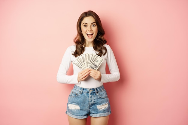 Free photo loans and microcredit. smiling beautiful girl showing money, cash in hands and looking enthusiastic, standing over pink background.