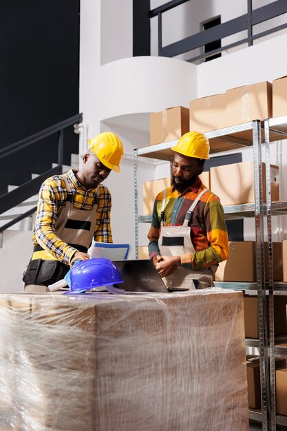 Free photo logistics managers analyzing inventory and comparing data on laptop and clipboard in warehouse. african american men checking goods supply schedule and working in distribution department storehouse