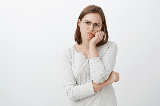 lonely displeased and unhappy cute young woman in glasses with short brown hair frowning and sulking from sadness leaning face on palm gazing with indifference over grey wall