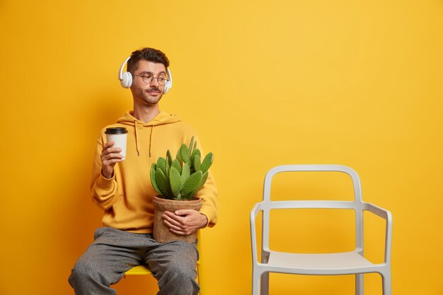 Lonely handsome guy spends free time alone holds potted cactus takeaway coffee looks at empty chair listens music via headphones