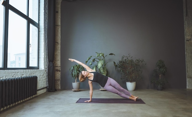 Одинокая женщина в спортивной одежде делает боковую планку в просторной студии йоги Спортивная женщина занимается хатха-йогой Полная длина Серый фон Фото высокого качества