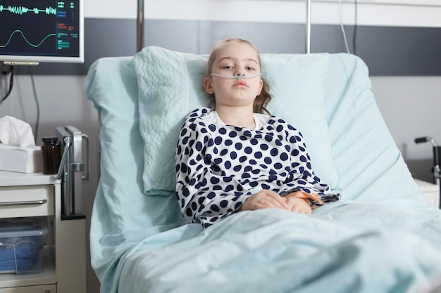 Lonely young pale sick girl patient resting in pediatric clinic bed while in recovery ward room. Ill child sitting alone in hospital bed while wearing nasal oxygen tube for life support.