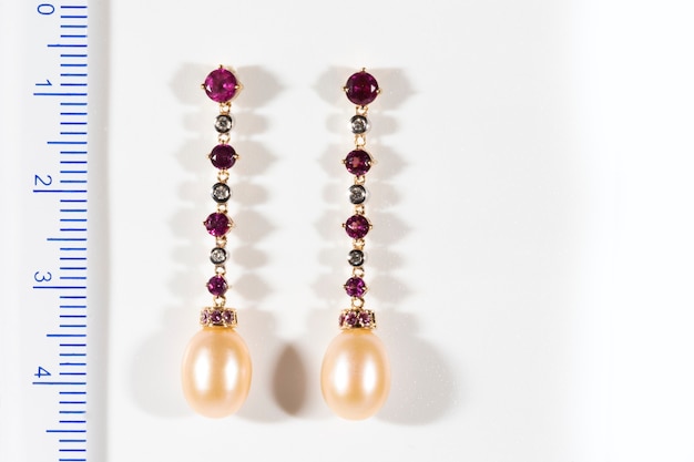 Long gold earrings with diamonds rubies and pearls on a white background next to the ruler