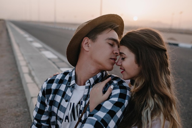 Free photo long-haired attractive girl gently embracing her boyfriend in stylish shirt with eyes closed on blur background. young man wearing hat going to kiss his girlfriend in nose walking across the road
