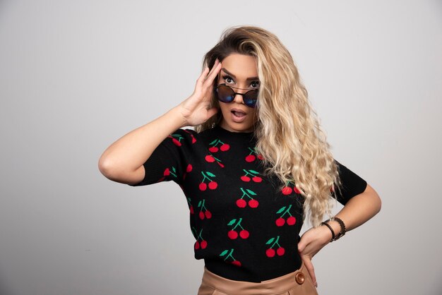 Long haired blonde woman in sun glasses standing on gray wall. 