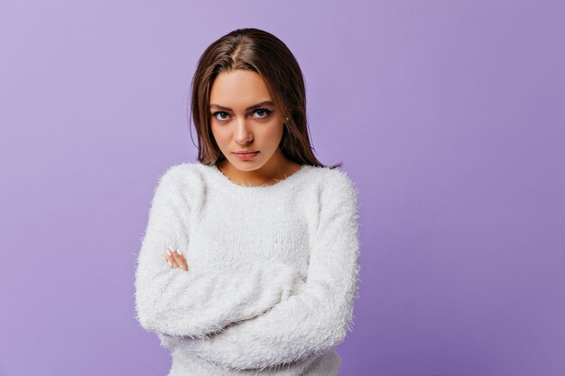 Long-haired brunette with green eyes offended and stands in closed pose. Closeup portrait of young girl in white warm sweater on lilac