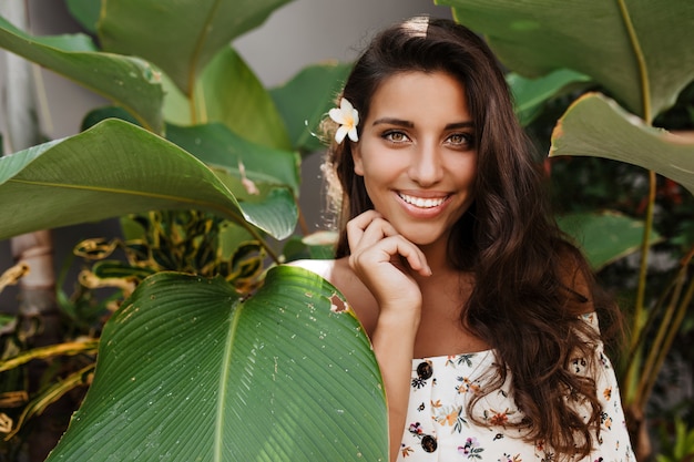 Long-haired brunette woman in great mood is posing with smile among tropical plants