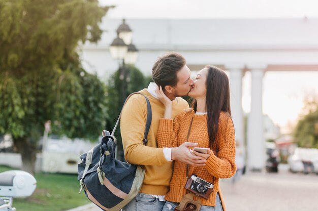 ぼやけた街で彼女のボーイフレンドにキスするカメラを持つ長い髪のブルネットの女性