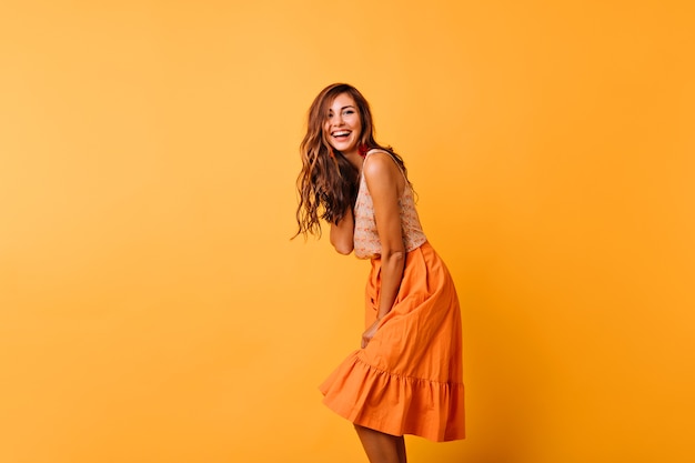 Длинноволосая кудрявая женщина в ярком наряде выражает положительные эмоции. Романтичная женская модель в оранжевой юбке, танцующей на желтом.