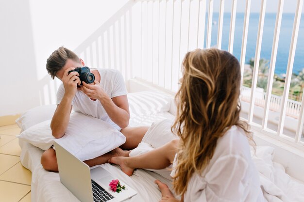 그녀의 남편이 사진을 만드는 동안 컴퓨터와 발코니에 쉬고 흰 셔츠에 긴 머리 곱슬 여자. 아침에 그의 화려한 아내의 사진을 찍는 전문 카메라를 가진 남자