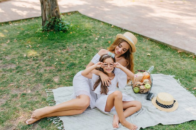 Long-haired cute girl came to park with her young mother to spend time together. Smiling woman in vintage hat looking at daughter which posing with peace sign, lying on blanket.