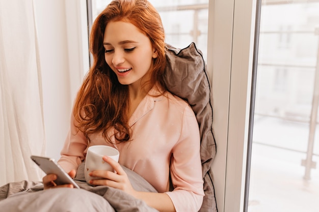 Long-haired ginger woman reading phone message in morning. Cute caucasian girl sitting in bed with cup of coffee and smartphone.