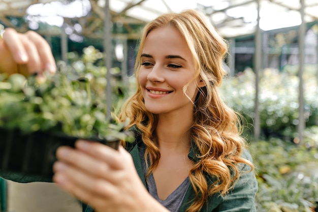 Long-haired redhead girl is happy and sincerely smiling, holding pot of greens in her hands. Closeup portrait outside surrounded by plants.