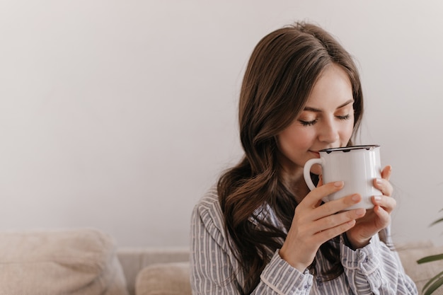 Long-haired woman in blue pajamas enjoying aroma of tea