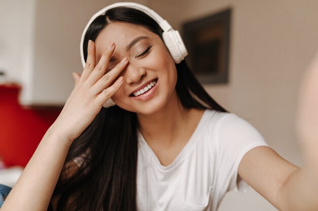 Long-haired woman in white top covers her face with her hand, laughs and takes selfie