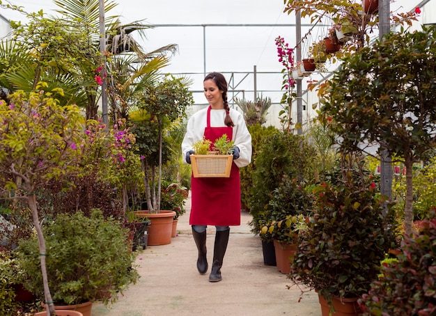 Long shot woman wearing gardening clothes and holding basket in greenhouse