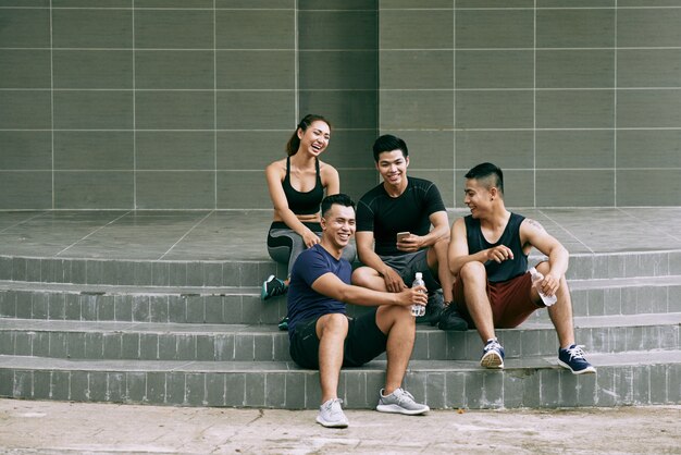 Long shot of young friends in sportswear resting on outdoor stairs and laughing joyfully