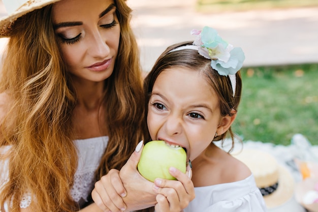 Lonkg-haired curly young woman with trendy makeup feeding daughter with green apple. Brunette little girl eating juicy fruit with big appetite during picnic in park.
