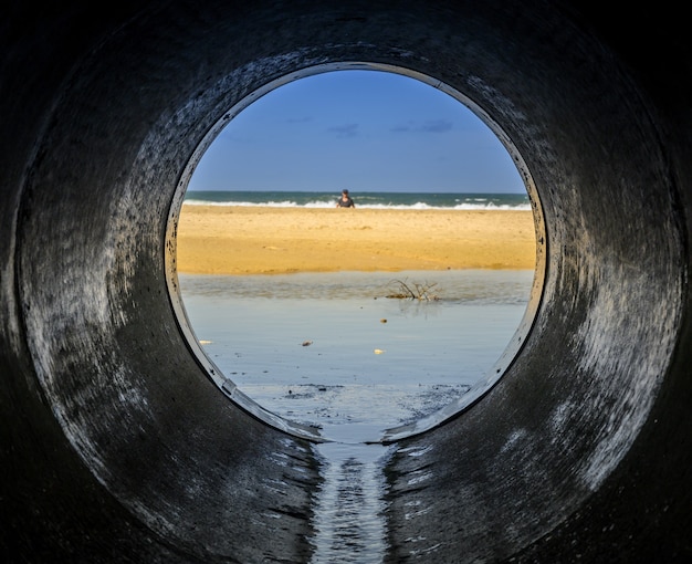 Free photo look form a pipe looking at the beach surrounded by the sea with people in it under sunlight