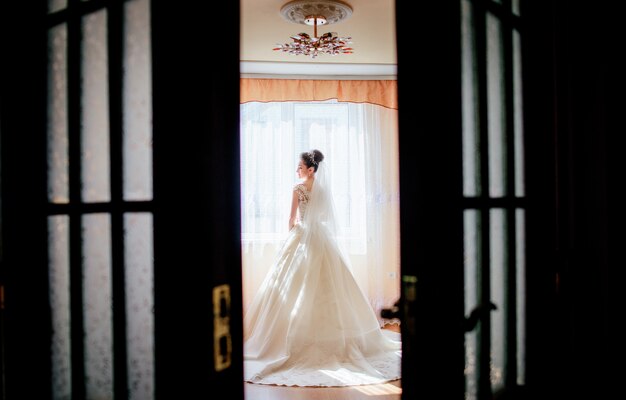 Look from behind the door at a pretty bride standing in a luxury hotel room