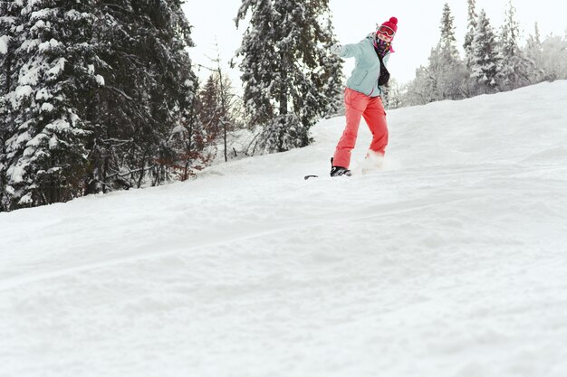 Look from below at woman in pink suit going down on the snowboard along the forest line 
