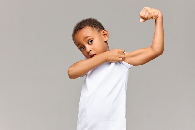 Look at my bicep. Picture of funny Afro American in casual white t-shirt posing isolated at gray wall pulling up sleeve, showing his tensed arm muscle. Childhood, fitness and sports concept