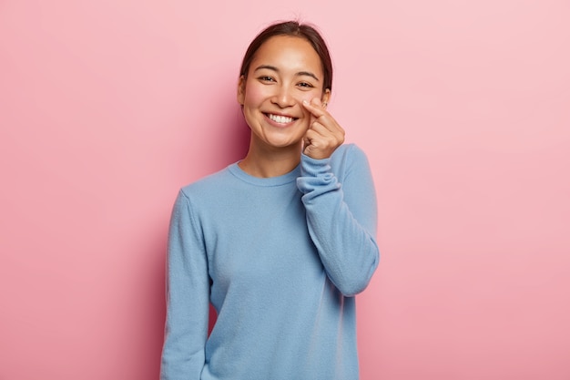 Look at my cheek. Pleasant looking Asian woman touches skin on face, demonstrates its freshness and softness, wears no make up, dark combed hair, dressed in casual blue sweatshirt, isolated on pink