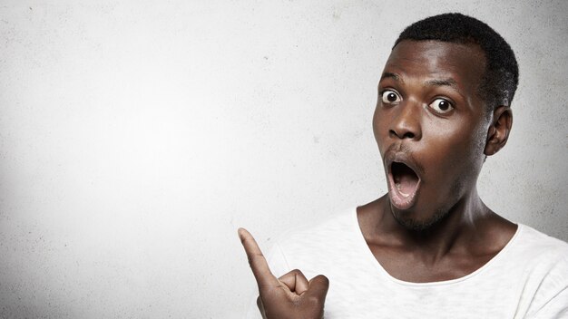 Look at that! Headshot of totally shocked bug-eyed African man dressed in white t-shirt, opening his mouth widely, pointing his finger at blank wall with copy space for your advertising content